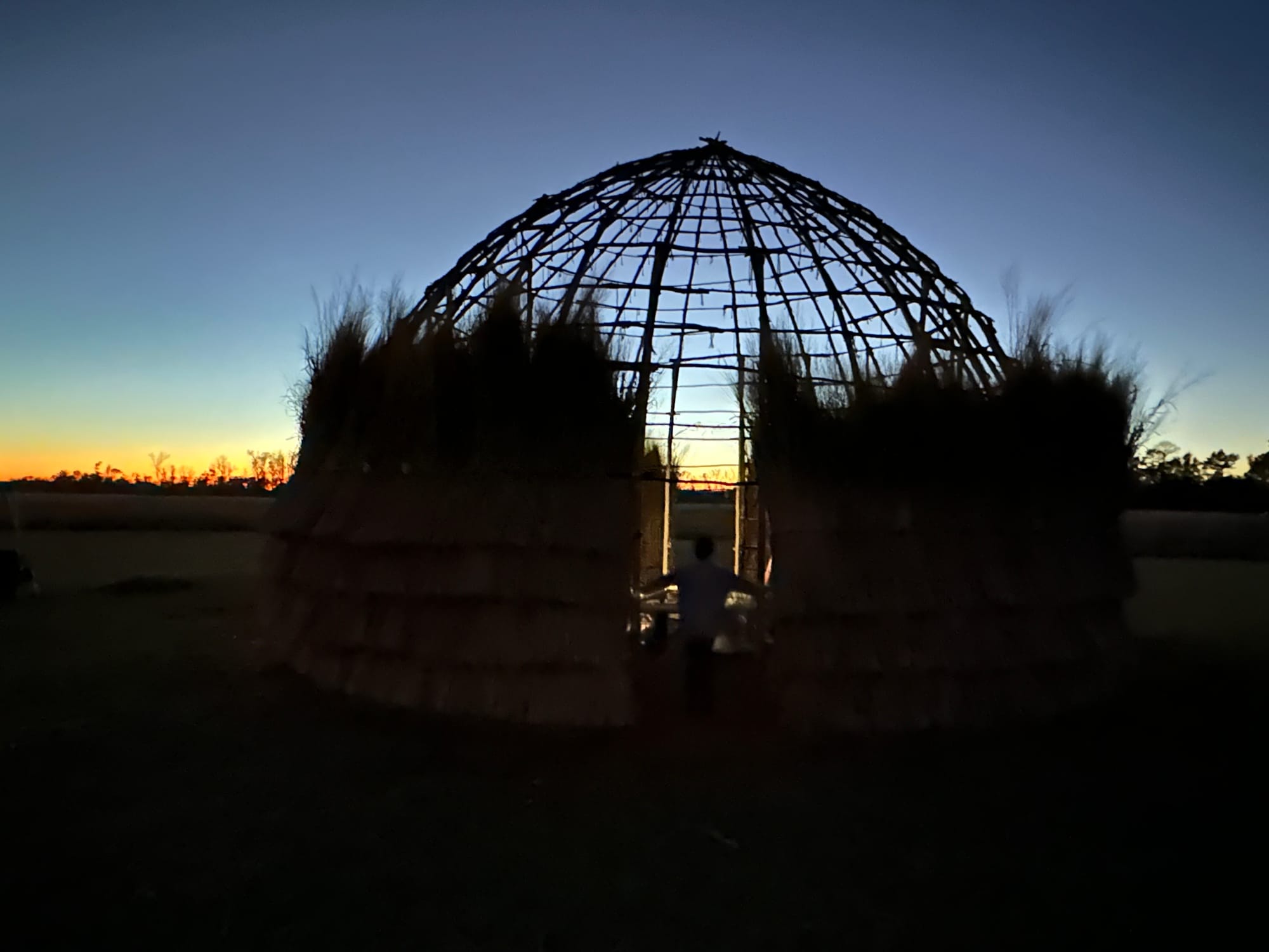 A Caddo grass house silhouetted against the sunset, its domed pole frame open to the sky